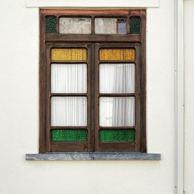 Janela de madeira com vidro colorido amarelo e verde e cortinas brancas numa parede branca e castanha