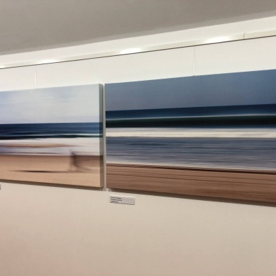 Dois quadros abstratos de paisagem costeira em tons de azul e castanho pendurados numa parede branca