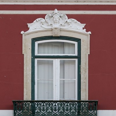 Janela de madeira branca com moldura verde e varandim de ferro forjado verde numa parede vermelha