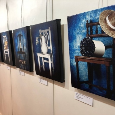 Conjunto de fotografias de Natureza Morta com cadeiras, jarros e chapéu em fundos azuis, penduradas numa parede creme