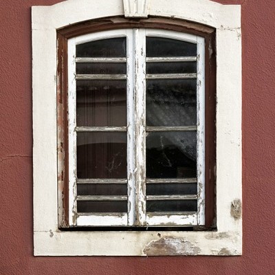 Janela de madeira antiga com persianas brancas desgastadas numa parede castanha avermelhada