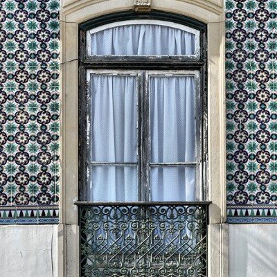 Janela com estrutura em madeira preta desgastada e grade de ferro ornamental em parede de azulejos coloridos