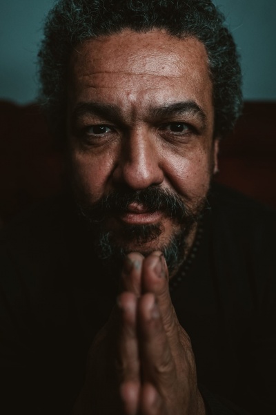 Homem com cabelo e barba grisalhos e mãos juntas em fundo escuro