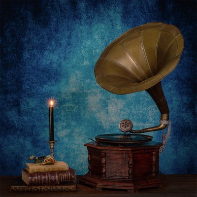 Gramofone antigo em madeira e latão ao lado de livros e vela acesa sobre mesa com fundo azul