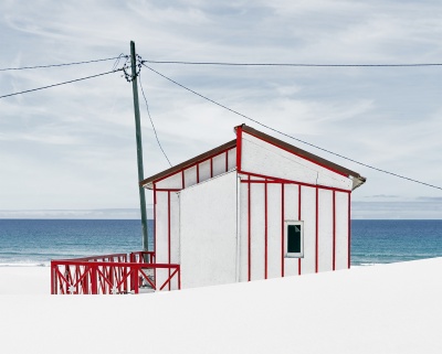 Casa pequena de praia branca com detalhes vermelhos rodeada por areia e mar ao fundo