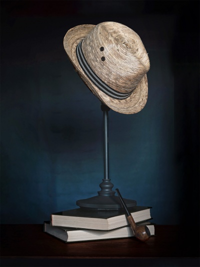 Chapéu de palha bege com faixa preta e branca em suporte preto, sobre livros e com cachimbo