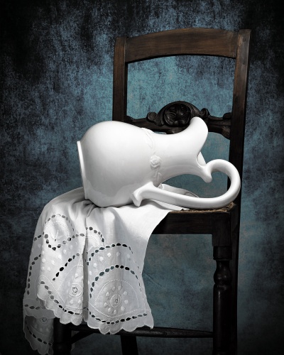 Jarra branca de cerâmica sobre toalha branca bordada em cadeira de madeira escura.