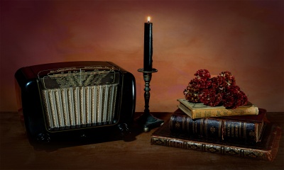 Rádio antigo, vela preta acesa num castiçal e livros antigos com flores secas numa mesa de madeira