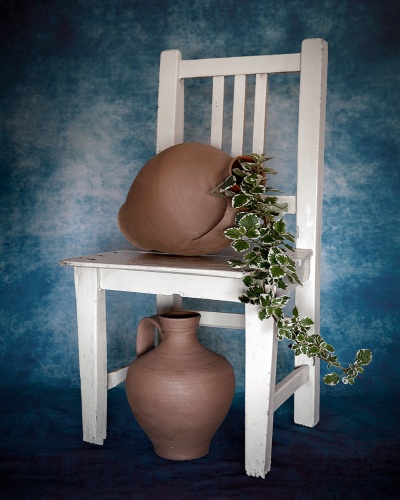Cadeira branca envelhecida com vaso de barro e planta, jarra de barro no chão, fundo azul.
