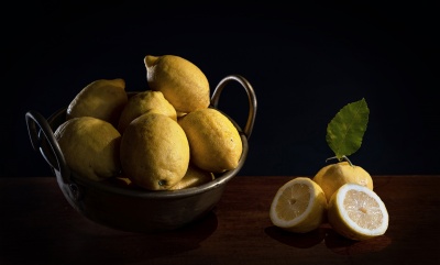 Cesto metálico com limas amarelas e lima cortada ao meio com folha num fundo escuro.