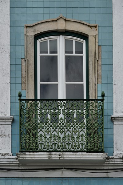 Janela branca dupla com varanda de ferro verde em fachada de azulejos azuis.