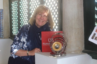 Mulher loira com óculos junto a escultura circular metálica em pedestal branco
