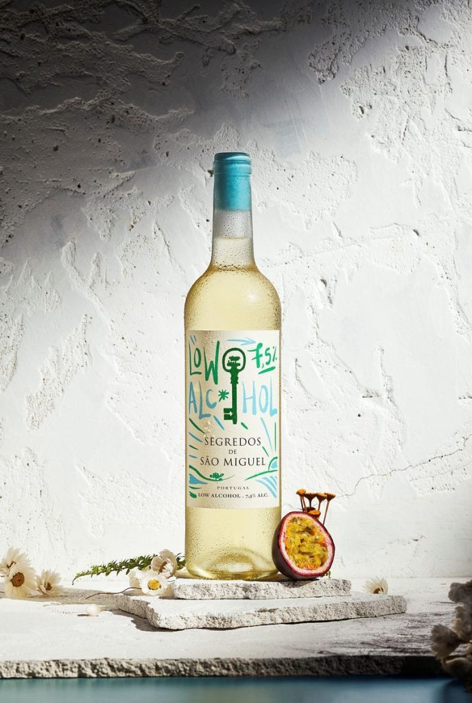 Segredos de São Miguel Branco LOW ALCOHOL Garrafa de vinho branco com rótulo azul e verde sobre pedra cinzenta com flores e fruto da paixão