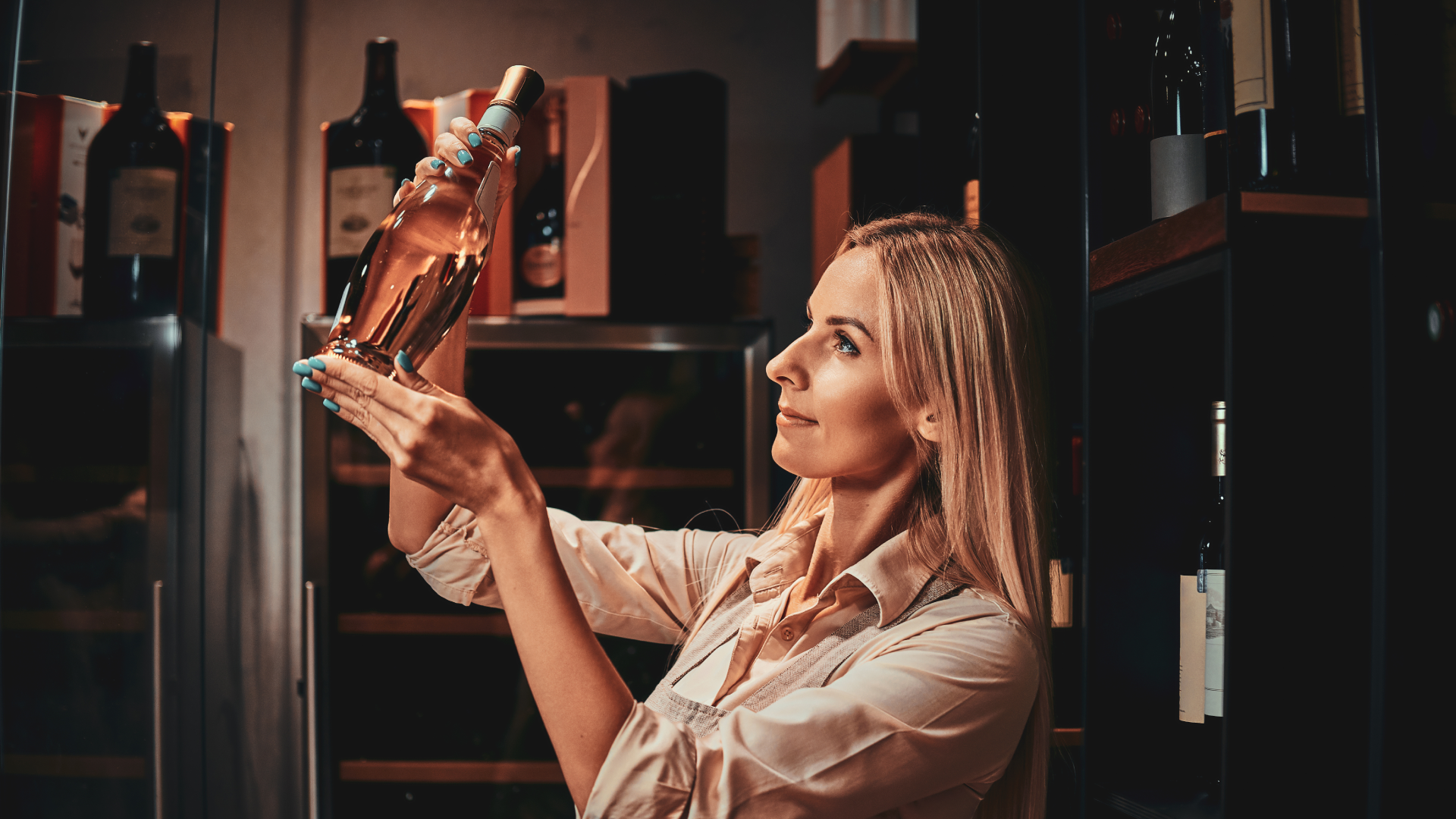 Mulher examinando uma garrafa de vinho rosé numa adega com prateleiras e outras garrafas