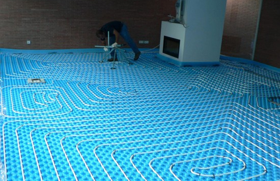 Sistema de aquecimento de piso com tubos brancos numa base azul em interior com paredes de tijolo.