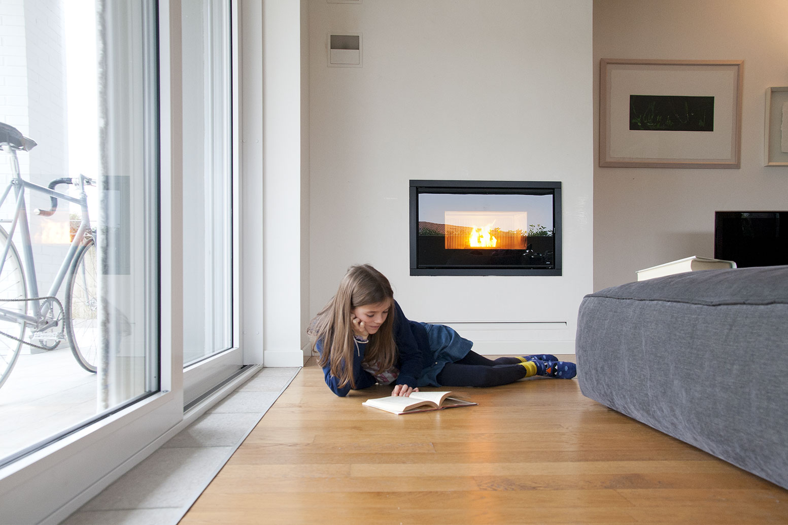 Menina deitada a ler no chão de madeira num interior com lareira e sofá cinzento