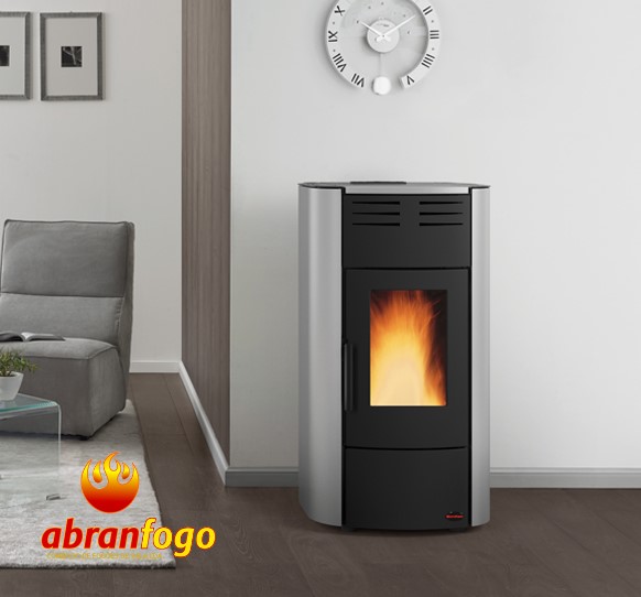 RAFFAELLA IDRO H15 Estufa de pellets metálica cinzenta com porta preta e fogo visível, com sofá cinzento e relógio na parede branca