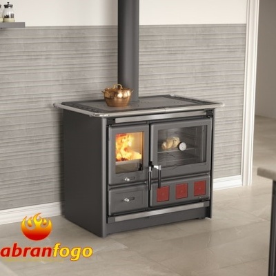 Fogão a lenha em aço negro com detalhes vermelhos e chaminé, em cozinha com parede cinza e chão claro