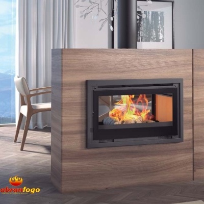 Lareira fechada embutida em madeira clara com fogo visível, cadeira clara e cortinas ao fundo