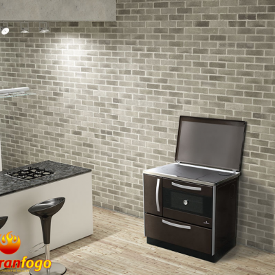 Fogão a lenha Abranfogo castanho escuro com tampo metálico prateado aberto numa cozinha moderna com parede de tijolo cinza e ilha branca.