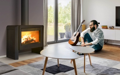 Lareira moderna preta acesa numa sala com mesa branca, tapete cinzento e homem sentado com guitarra