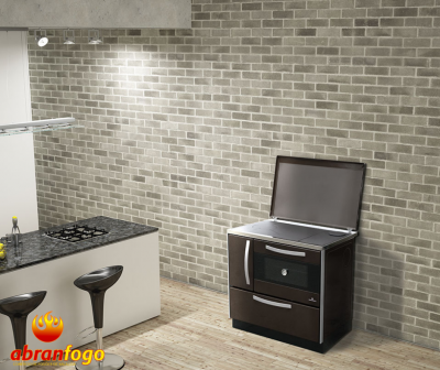 Fogão a lenha Abranfogo castanho escuro com tampo metálico prateado aberto numa cozinha moderna com parede de tijolo cinza e ilha branca.