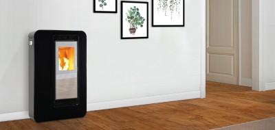 Estufa de pellets preta com vidro frontal mostrando fogo, em chão de madeira e parede branca decorada com quadros de plantas