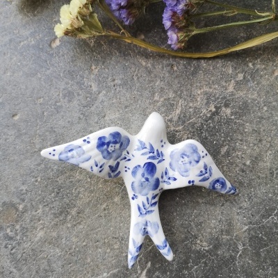 Pássaro decorativo em cerâmica branca com padrão azul de flores sobre superfície cinza com flores secas.