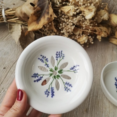 Prato cerâmico branco com flores pintadas em roxo e verde e recortes florais, com fundo de flores secas e folhas.