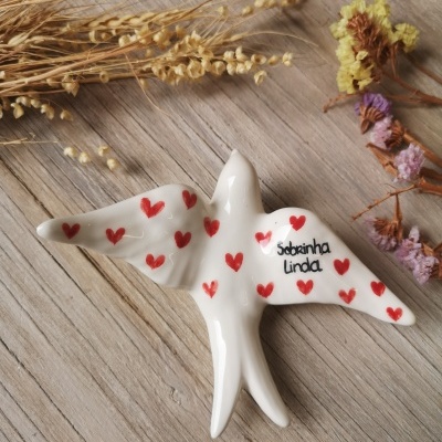 Pássaro decorativo em cerâmica branca com corações vermelhos e texto sobre superfície de madeira com flores secas