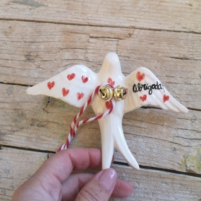 Pomba de cerâmica branca com corações vermelhos e texto Obrigada, com guizos dourados, segurada por mão feminina