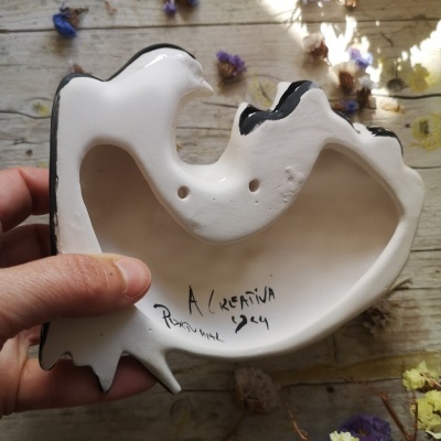 Cinzeiro de cerâmica branco com detalhes a preto e assinaturas em superfície de madeira com flores secas.