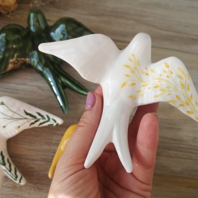 Pássaros decorativos em cerâmica, branco com detalhes dourados, branco com verde, verde e amarelo, sobre superfície de madeira.