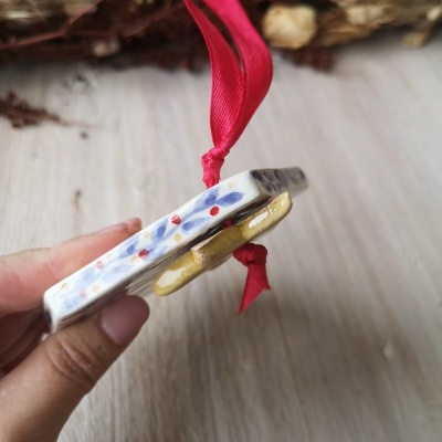 Ornamento de cerâmica com padrão de folhas e bolinhas e fita vermelha para pendurar