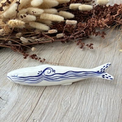 Figura de peixe decorativa branca com padrão azul sobre mesa de madeira com flores secas ao fundo
