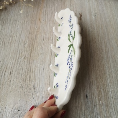 Suporte de cerâmica branco com padrão floral azul e verde e ganchos, com fundo de madeira e flores secas.