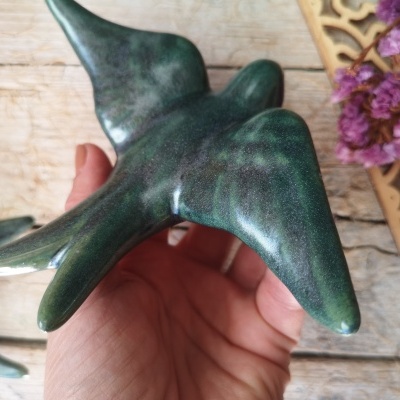 Escultura em cerâmica verde de pássaro segurada por mão sobre madeira com flores ao fundo