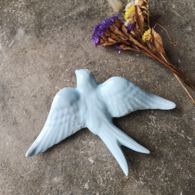Figura branca de pássaro em cerâmica com flores secas ao lado sobre superfície de pedra