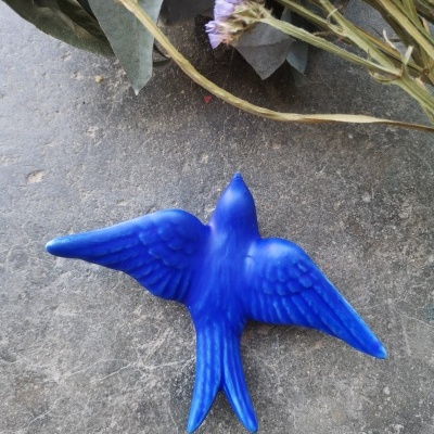 Pássaro decorativo azul com asas abertas sobre superfície cinzenta com flores secas ao fundo