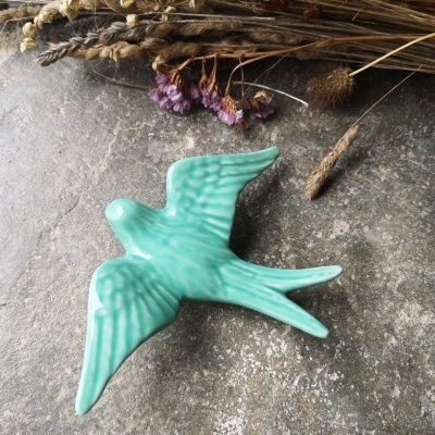 Estatueta em cerâmica verde de andorinha sobre superfície cinza com flores secas