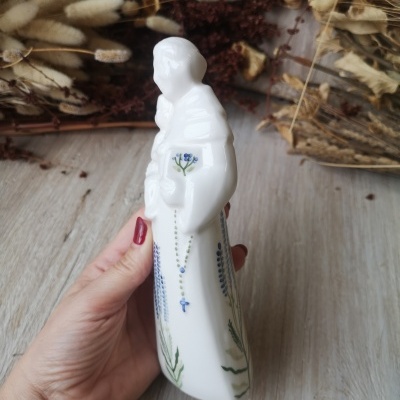 Figura religiosa de porcelana branca com pintura floral azul e verde e rosário azul, segurada por mão com unhas vermelhas.