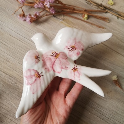 Estatueta cerâmica branca pássaro com flores cor-de-rosa em mão, fundo madeira clara e flores secas
