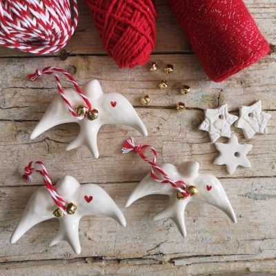 Enfeites de Natal em forma de andorinha em cerâmica branca com corações vermelhos e sinos dourados, fios vermelhos e brancos e estrelas brancas sobre madeira rústica