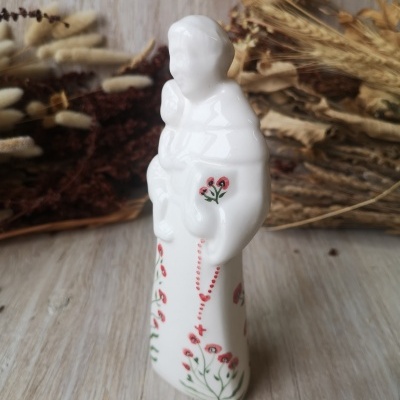 Figura de cerâmica branca com flores pintadas e rosário, sobre madeira e ramos secos.