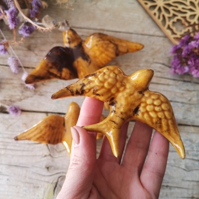 Três pássaros de cerâmica castanha com detalhes pretos, um na mão, sobre fundo de madeira clara com flores roxas secas e objeto decorativo de madeira