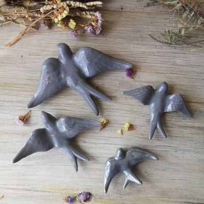 Quatro figuras de pássaros cinzentos em cerâmica numa mesa de madeira com ramos e flores secas