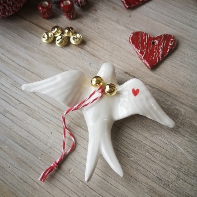 Pássaro de cerâmica branca com coração vermelho e sinos dourados sobre madeira clara