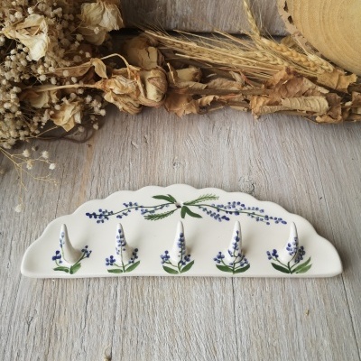 Gancho cerâmico branco decorado com flores lilás e folhas verdes sobre madeira clara com flores secas ao fundo.