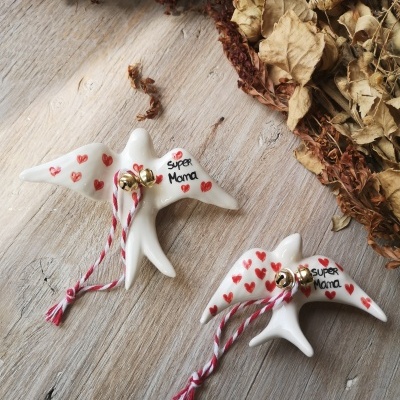 Dois pássaros cerâmicos brancos com corações vermelhos e cordões com sinos dourados, sobre madeira clara e folhas secas