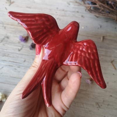 Escultura vermelha de cerâmica em forma de pássaro com asas abertas, apoiada numa mão.